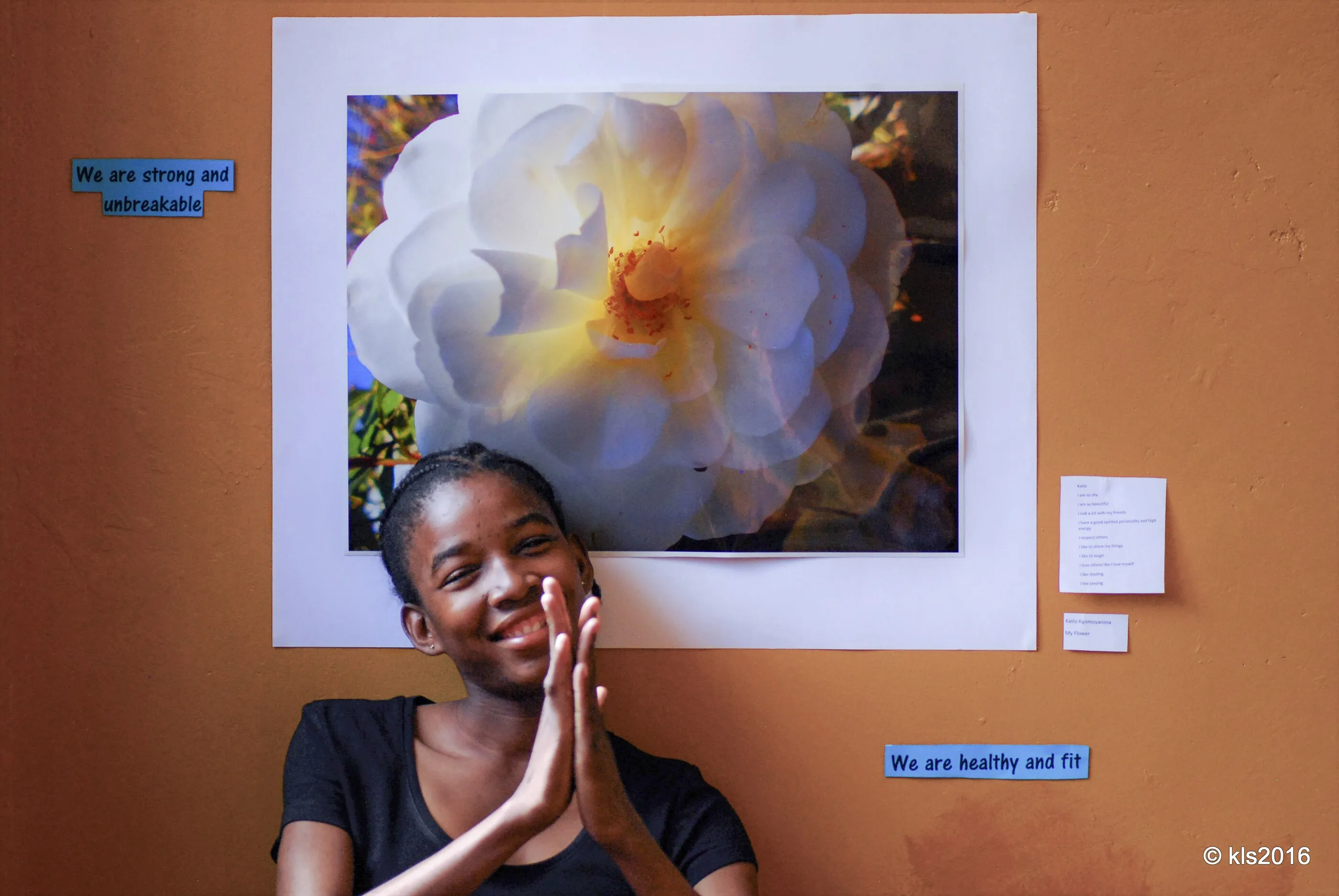 A young woman in Botswana smiling beside her exhibited photograph of a white flower, with the words 'We are strong and unbreakable' on the wall