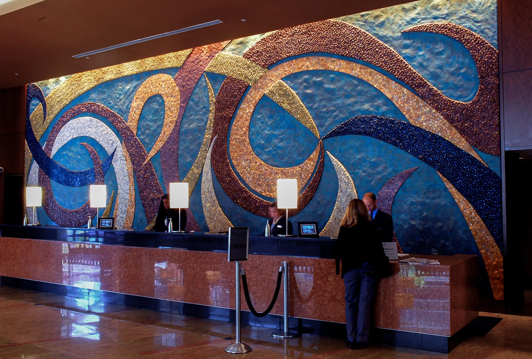 Conversing Waters, a sweeping stone mosaic and plaster mural of blue, copper and gold curves behind a hotel reception desk