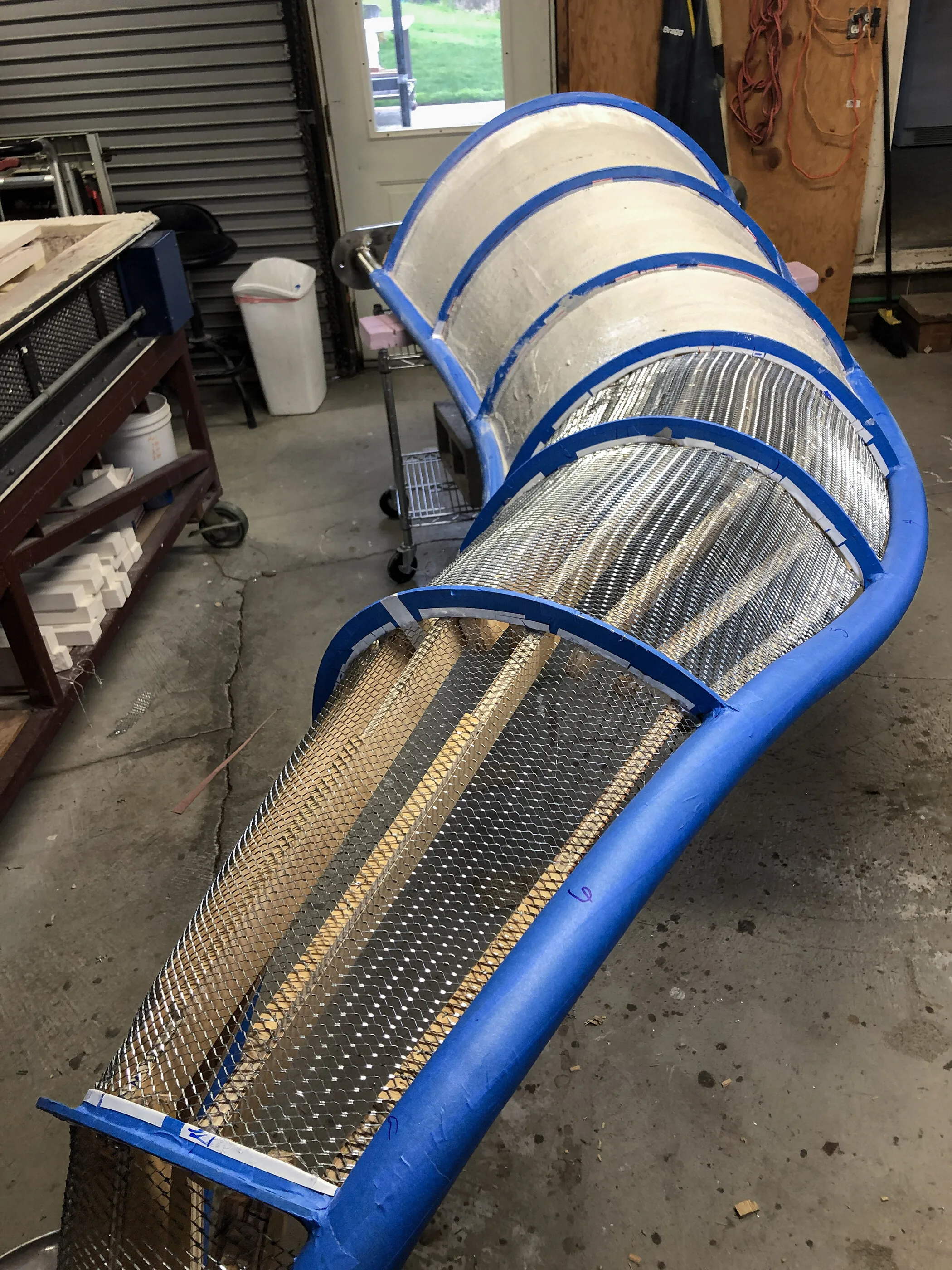 The steel armature of River Waltz in the studio, a curved blue frame with a mesh skin, before the glass goes on