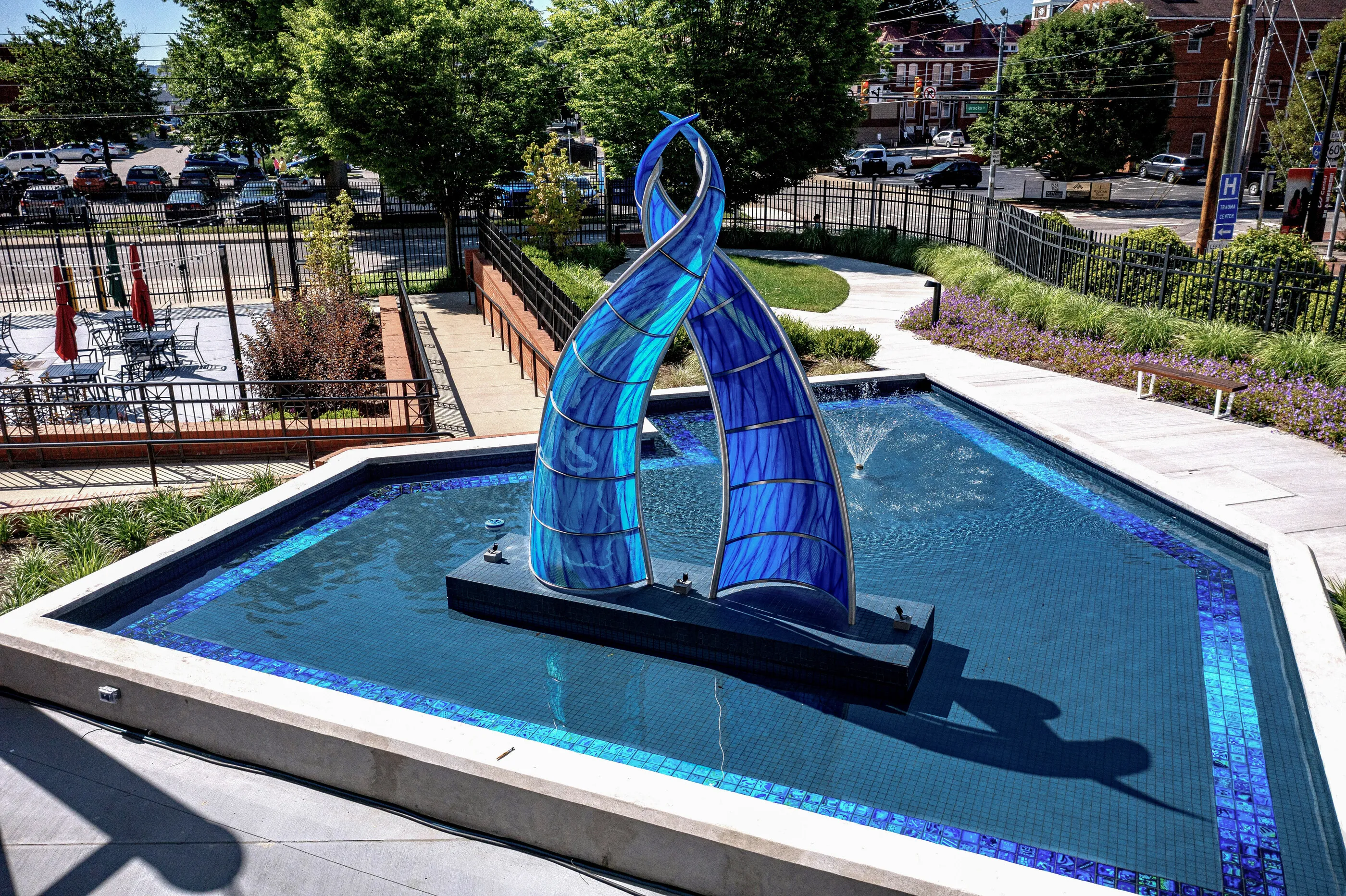River Waltz installed: two twisting blue glass and steel forms rising from a reflecting pool lined with blue tile, with the city behind